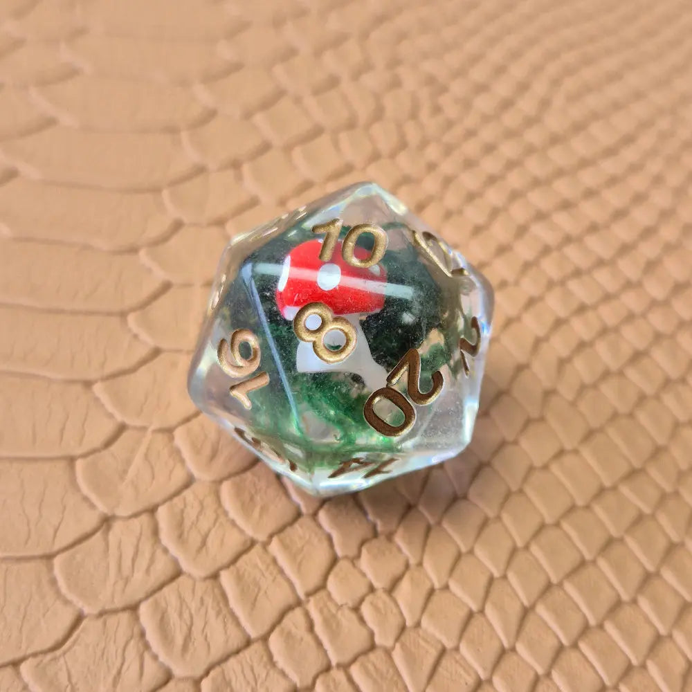 Clear resin D20 with red mushroom and moss encased inside, golden numbers, set on a textured leather background.